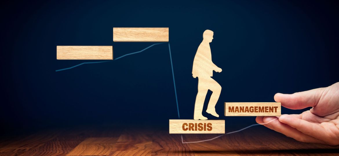 Person stepping from a block labeled "CRISIS" to a block labeled "MANAGEMENT," with a hand supporting the "MANAGEMENT" block.