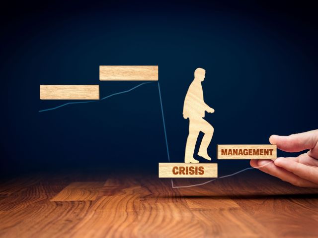 Person stepping from a block labeled "CRISIS" to a block labeled "MANAGEMENT," with a hand supporting the "MANAGEMENT" block.